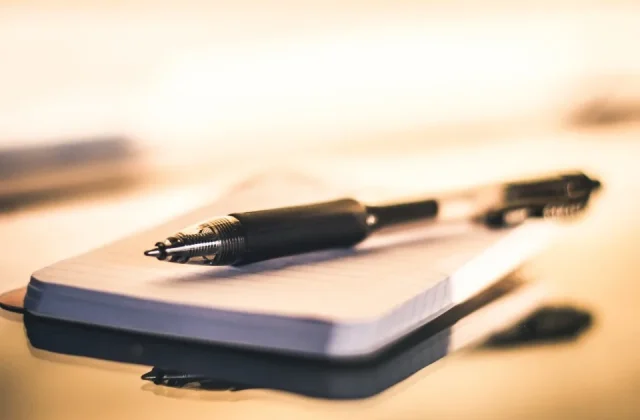 a hand writing notes in a notebook on a desk beside a laptop