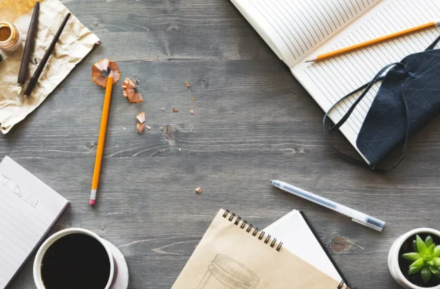 a hand writing notes in a notebook on a desk beside a laptop