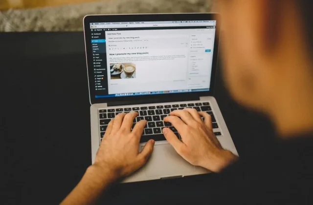 hands typing on a keyboard beside a laptop and a screen