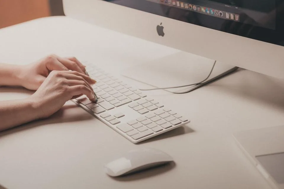 hands typing on a keyboard beside a laptop and a screen