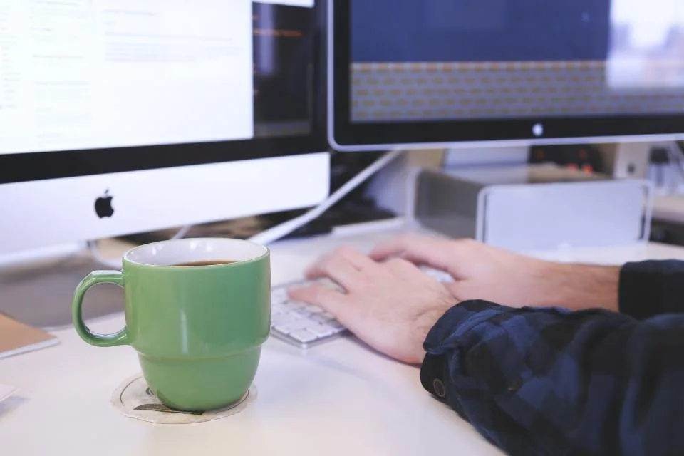 hands typing on a keyboard beside a laptop and a screen
