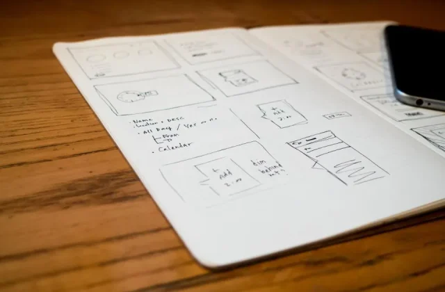 a page of interface sketches on paper beside a laptop and a pen
