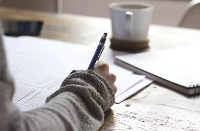 a hand writing notes in a notebook on a desk beside a laptop