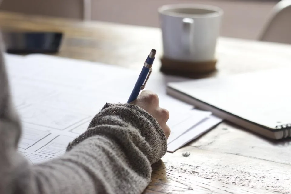 a hand writing notes in a notebook on a desk beside a laptop