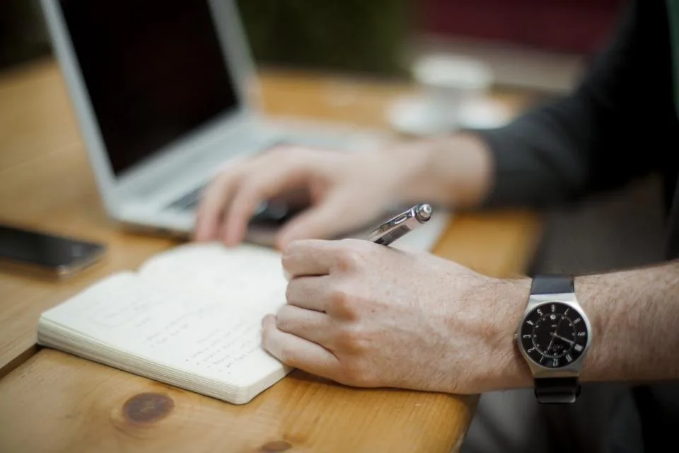 a hand writing notes in a notebook on a desk beside a laptop