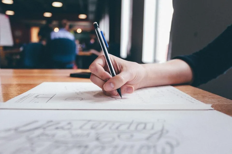 a hand writing notes in a notebook on a desk beside a laptop