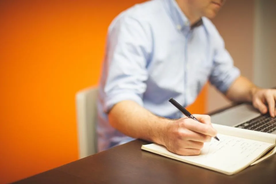 a hand writing notes in a notebook on a desk beside a laptop