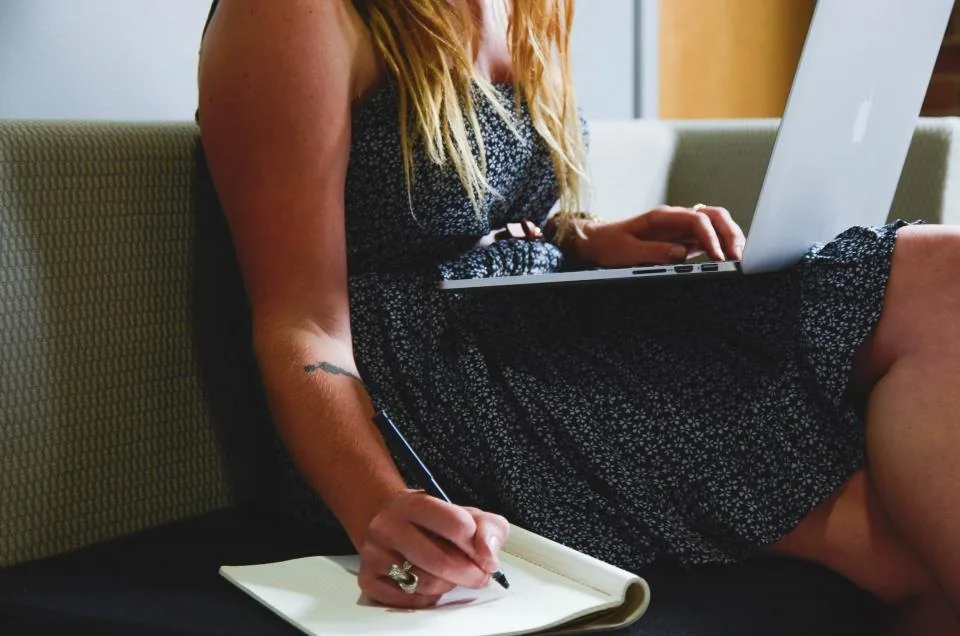 a hand writing notes in a notebook on a desk beside a laptop