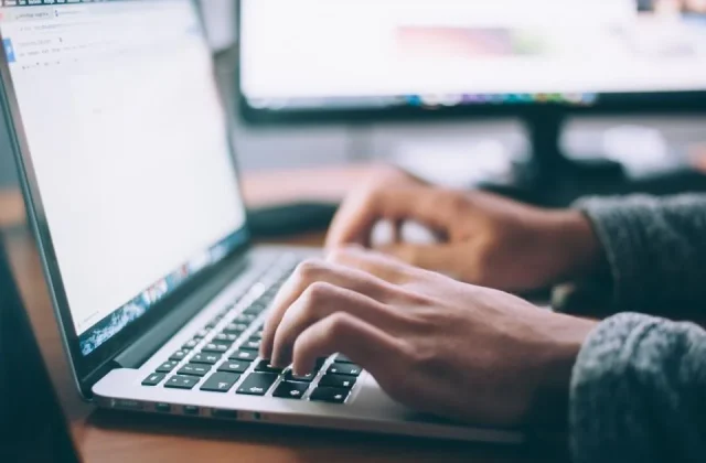 hands typing on a keyboard beside a laptop and a screen