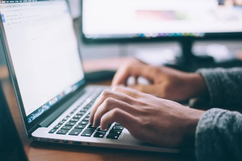 hands typing on a keyboard beside a laptop and a screen
