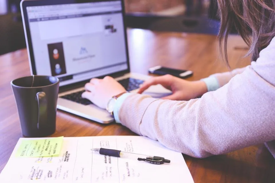 a laptop open on a desk beside a notebook, a phone and a cup of coffee