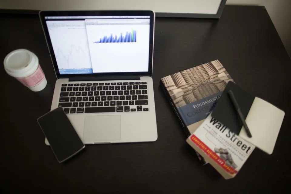 a laptop open on a desk beside a notebook, a phone and a cup of coffee