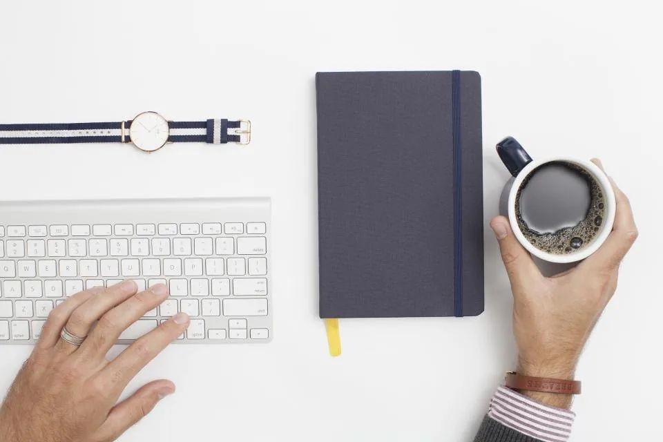 a close view of a desk with a keyboard, a notebook, a pen and a coffee cup