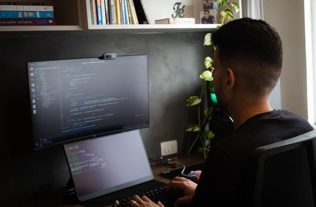 A developer writing code on a laptop with a large monitor showing the same project beside it