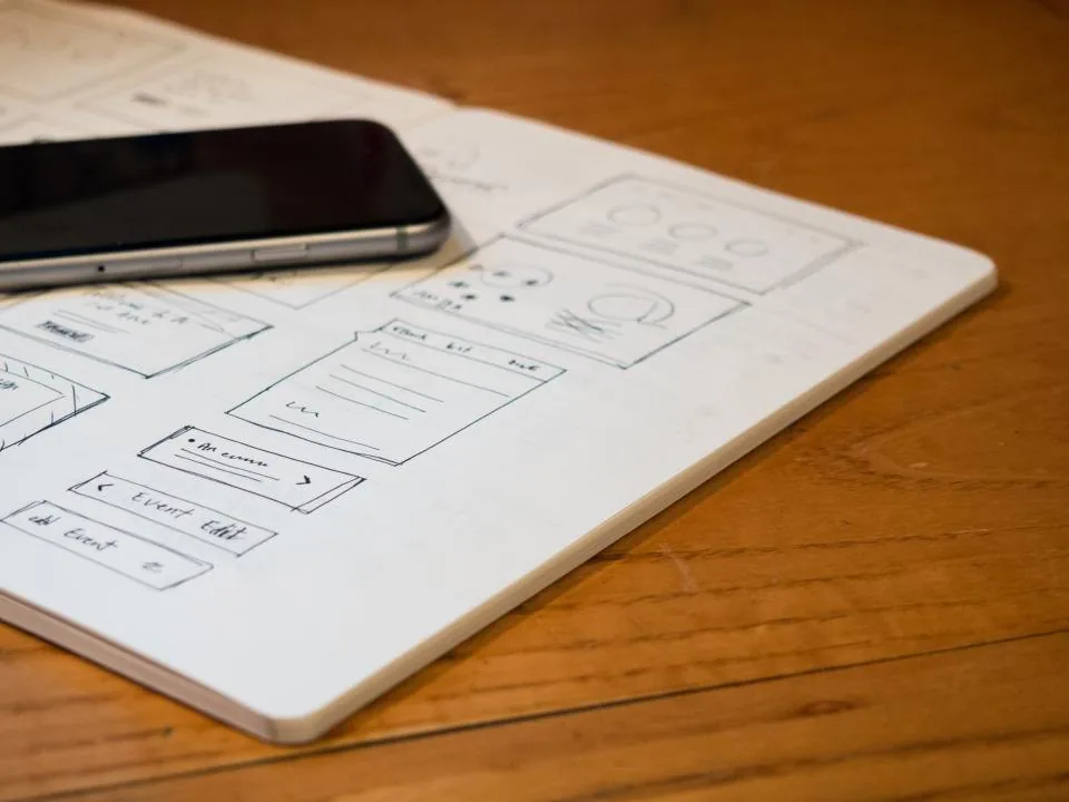a page of interface sketches on paper beside a laptop and a pen