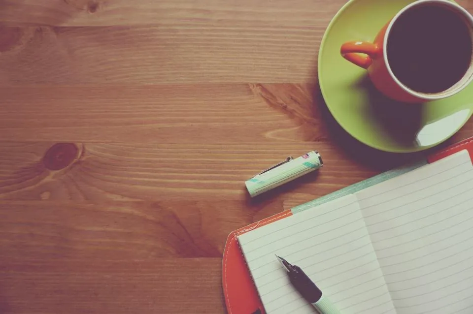 a hand writing notes in a notebook on a desk beside a laptop
