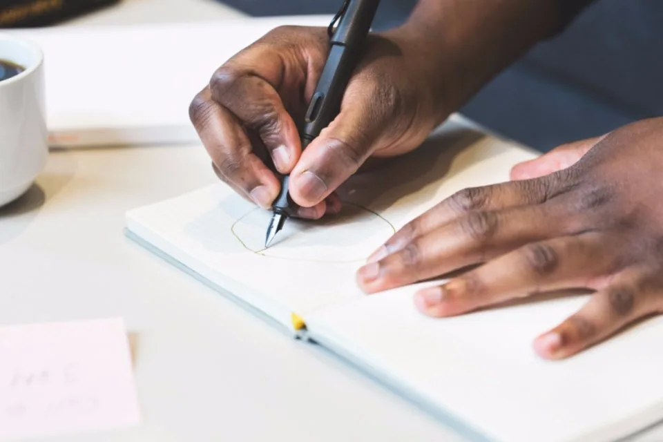 a hand writing notes in a notebook on a desk beside a laptop