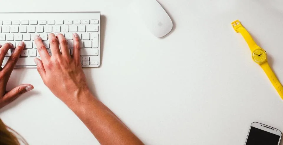 hands typing on a keyboard beside a laptop and a screen