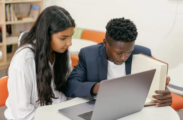 A teacher and a student reading a book together at a desk.